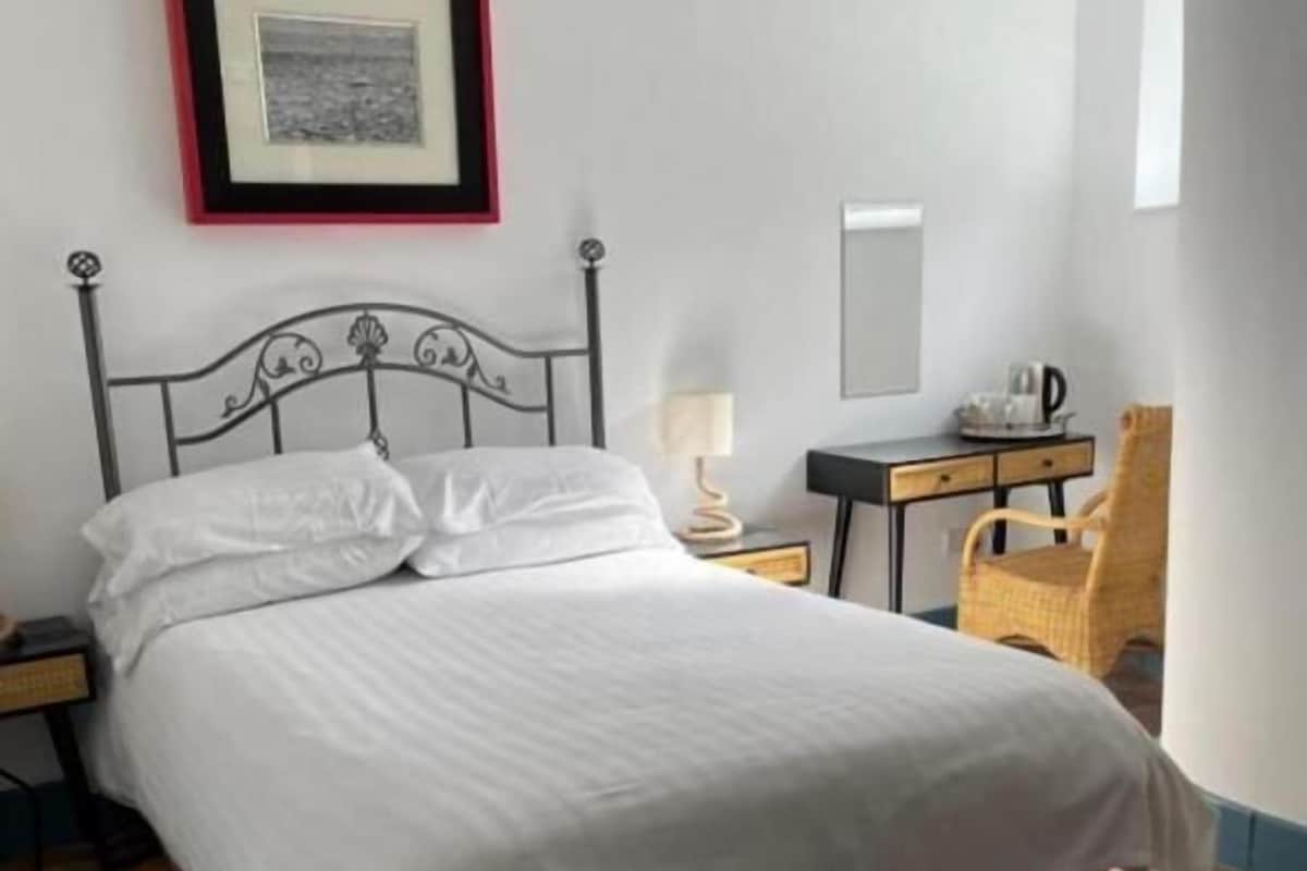 A neatly made bed with a white comforter, a bedside table with a lamp, a chair, and a framed picture on the wall.