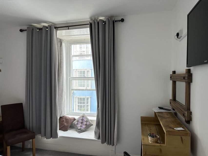 A room with a window, a chair, a wooden desk, and a television mounted on the wall.