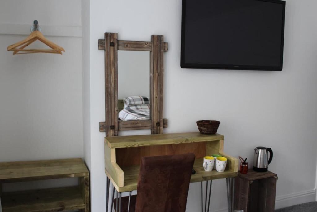Ein Zimmer mit Schreibtisch aus Holz, einem Stuhl, einem Spiegel, einer Tafel und einem kleinen Regal mit Tassen und Wasserkocher.