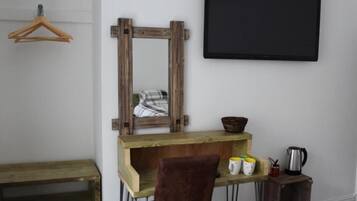 Ein Zimmer mit Schreibtisch aus Holz, einem Stuhl, einem Spiegel, einer Tafel und einem kleinen Regal mit Tassen und Wasserkocher.