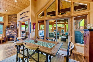 A spacious room with a large wooden table, chairs, a stone fireplace, and large windows offering views of the outdoors. - Newly Listed Breckenridge Riverfront Retreat with Mountain View’s (Breckenridge)