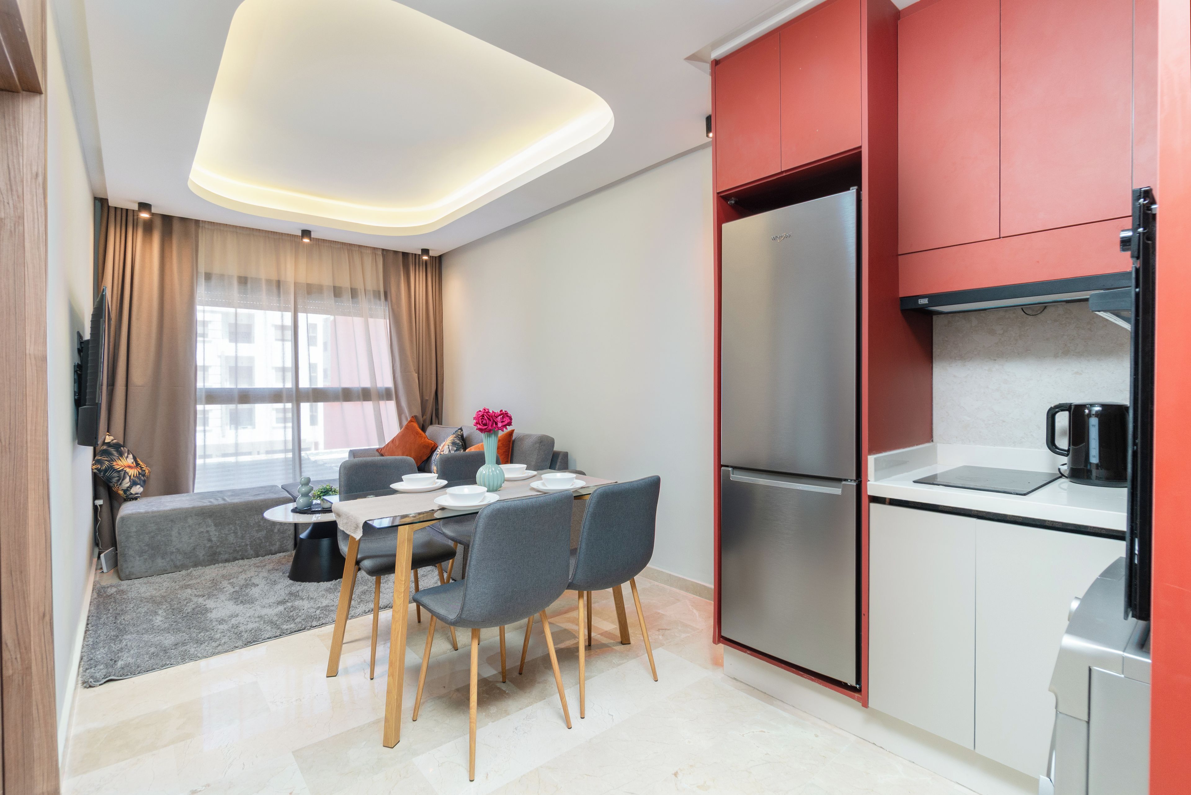 A modern kitchen with a stainless steel refrigerator, a dining area with a table and chairs, and a living space with a sofa and a window with curtains.
