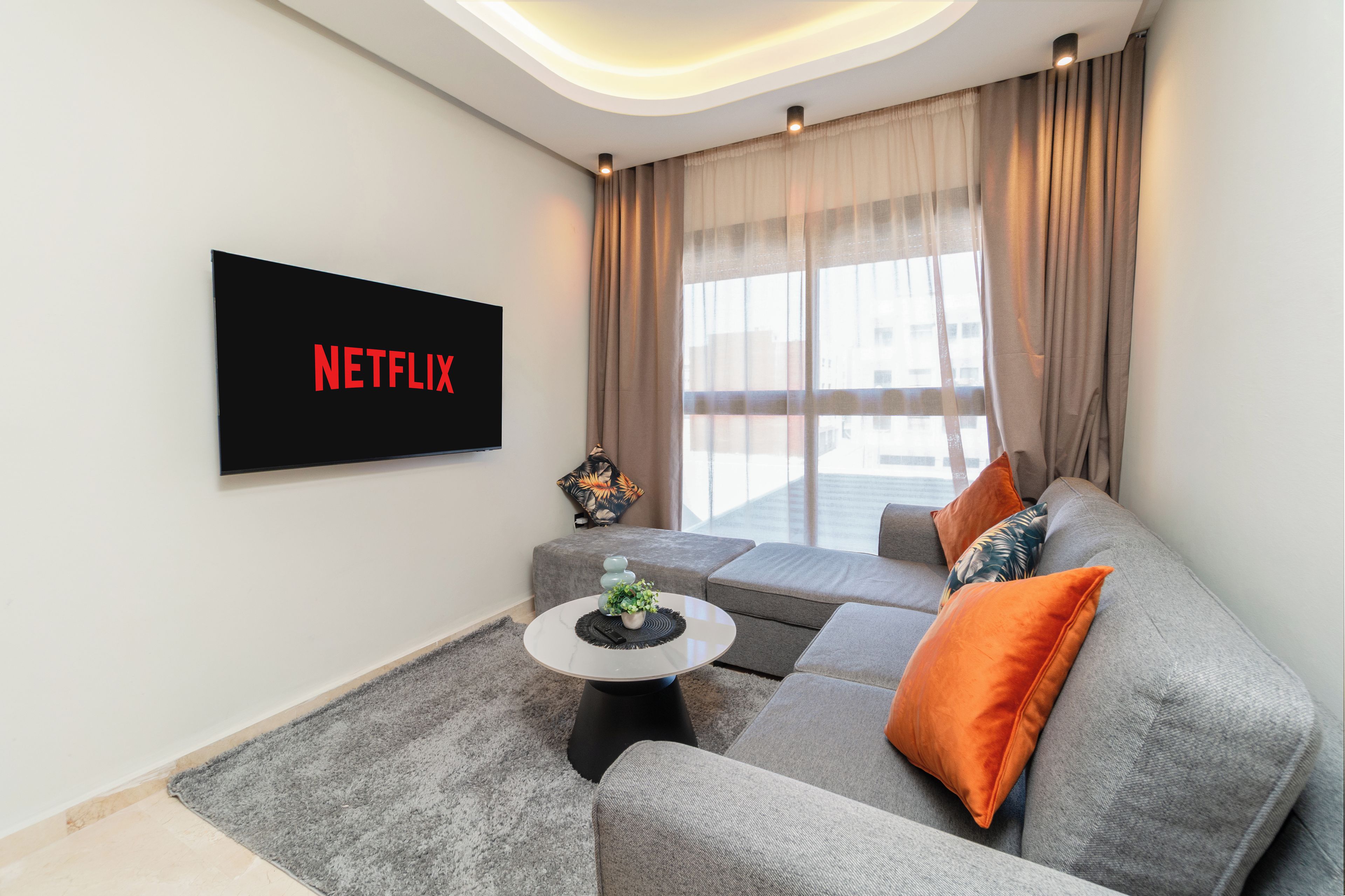 A modern living room with a grey sofa, a coffee table, and a flat-screen TV mounted on the wall displaying 'NETFLIX'.