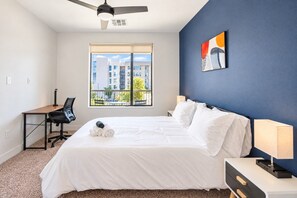Ein Hotelzimmer mit einem Bett, einem Schreibtisch, einem Sessel, einem Deckenventilator, einem Fenster mit Blick auf Gebäude und einer Wand mit einem Gemälde.