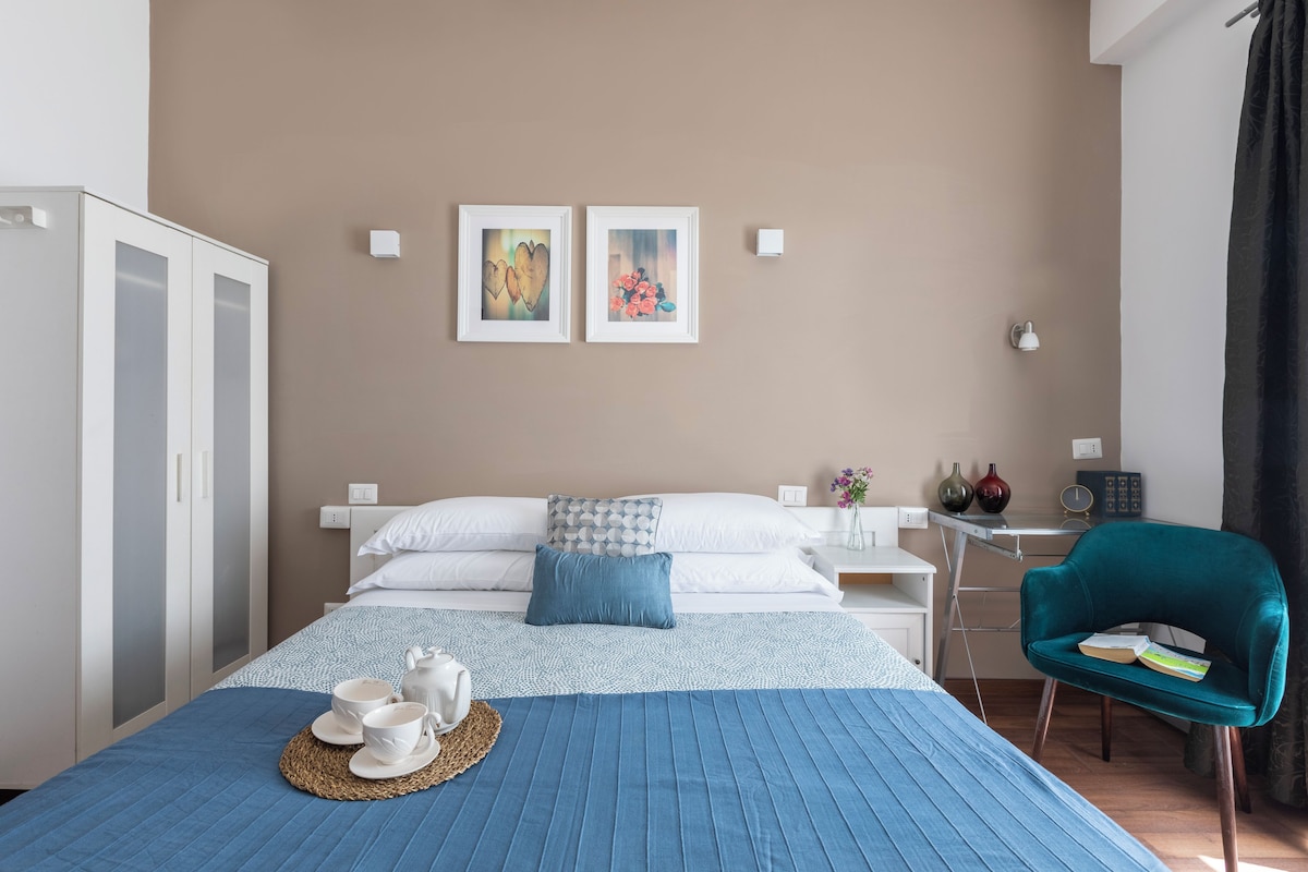 A hotel room with a bed, bedside tables, a desk, a chair, and artwork on the wall.