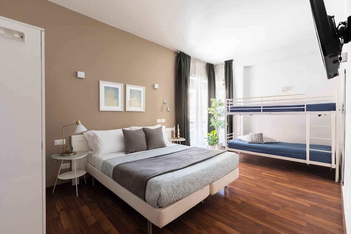 A bedroom with a bunk bed, a bedside table with a lamp, a small bedside table with a clock, and a wall-mounted light.