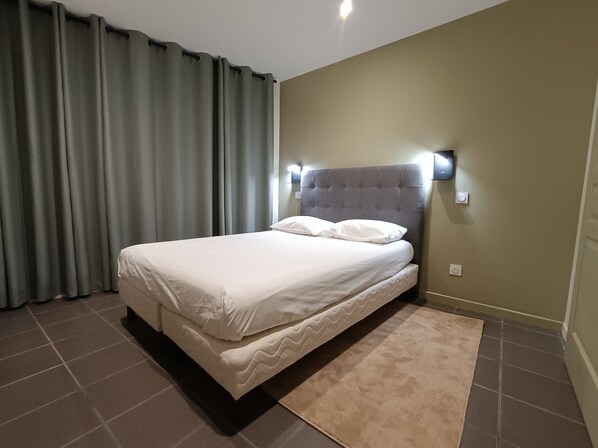 A hotel room with a bed, curtains, a headboard with wall-mounted lights, and a carpet.