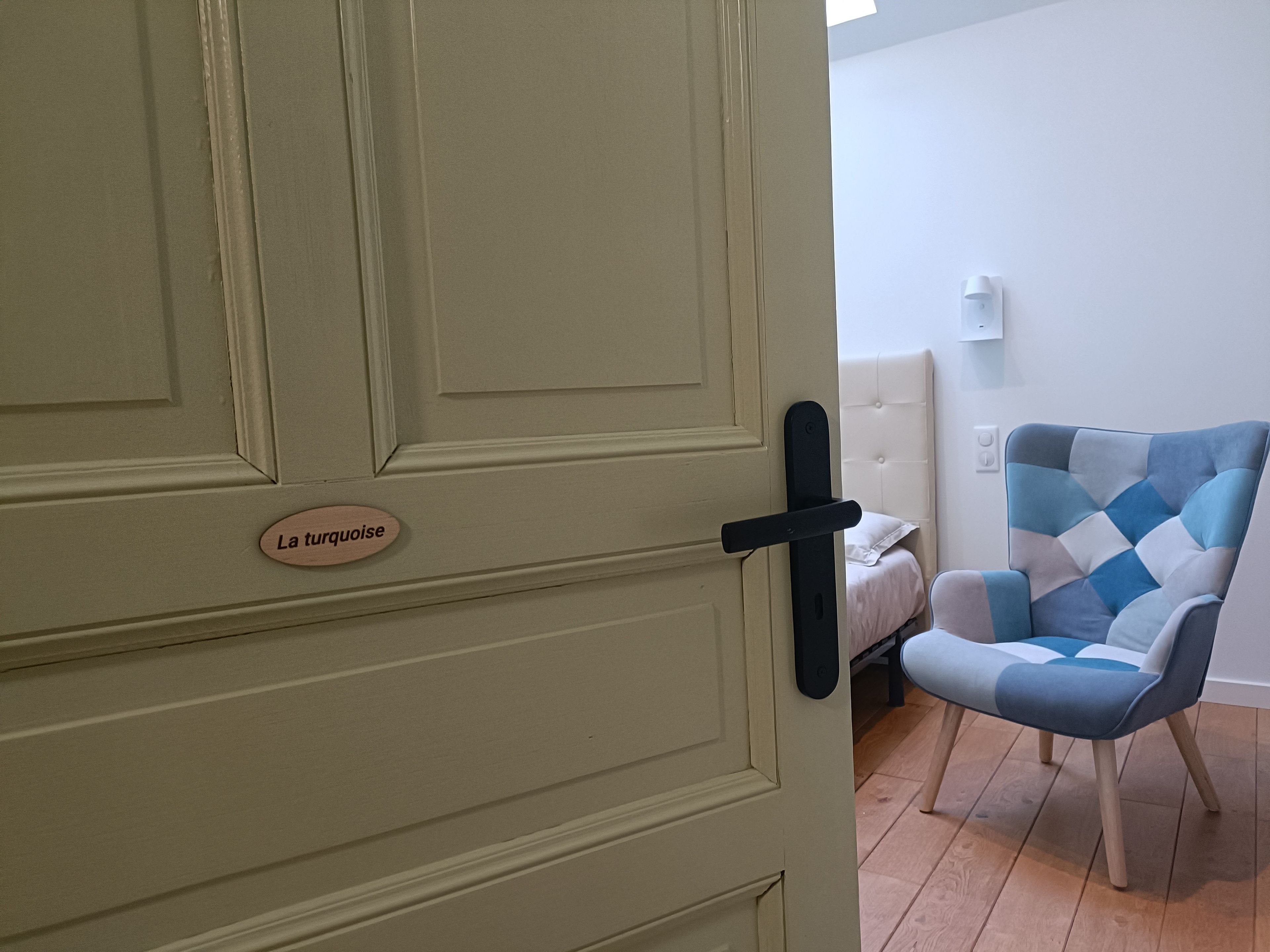 Una stanza con una porta con la scritta 'La turquoise', un letto con testiera bianca e una poltrona azzurra e bianca a motivi decorativi.