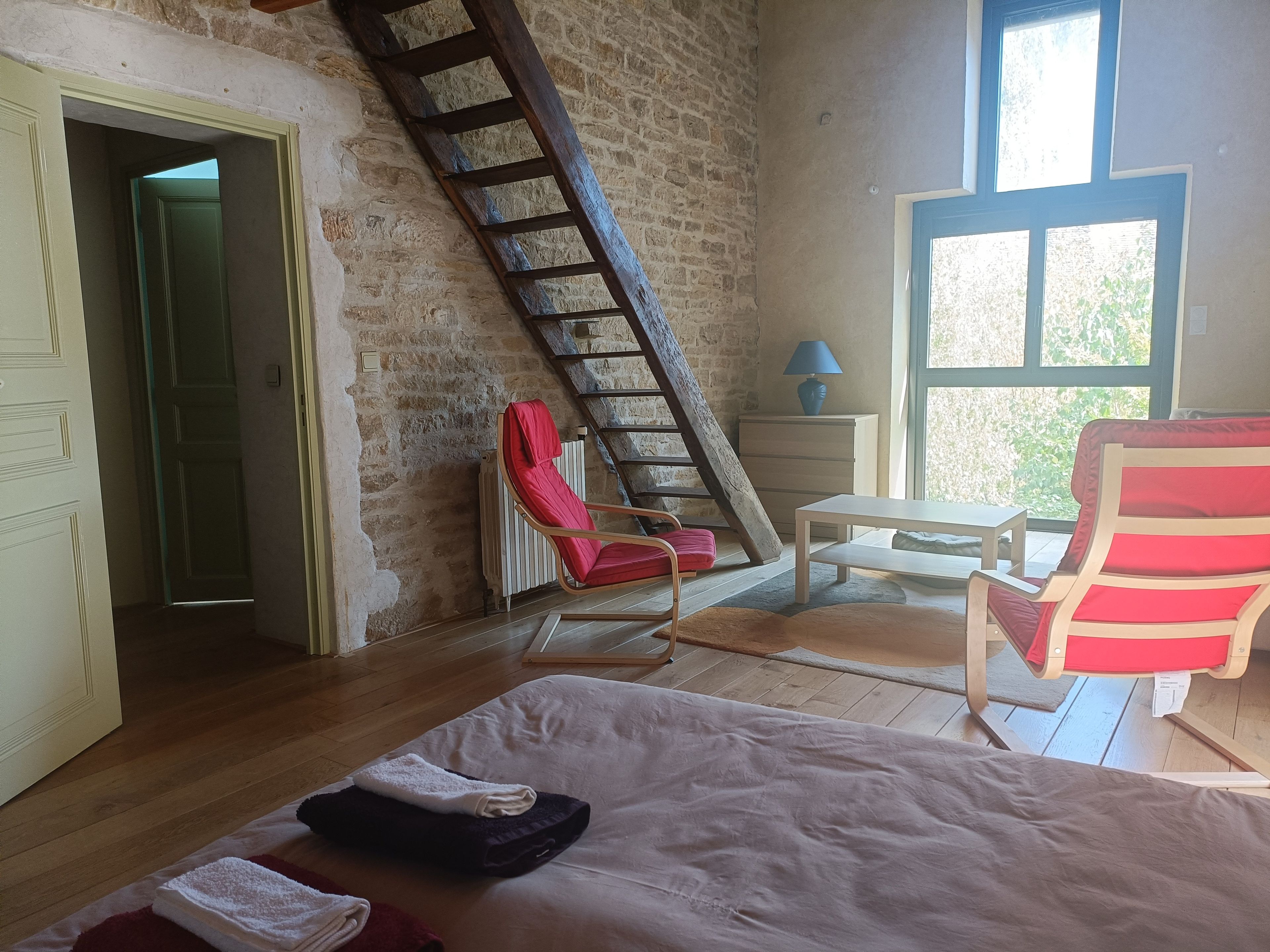 A room with a wooden floor, a bed with a white and brown cover, a red chair, a wooden table, a blue lamp, and a window with a view of greenery.