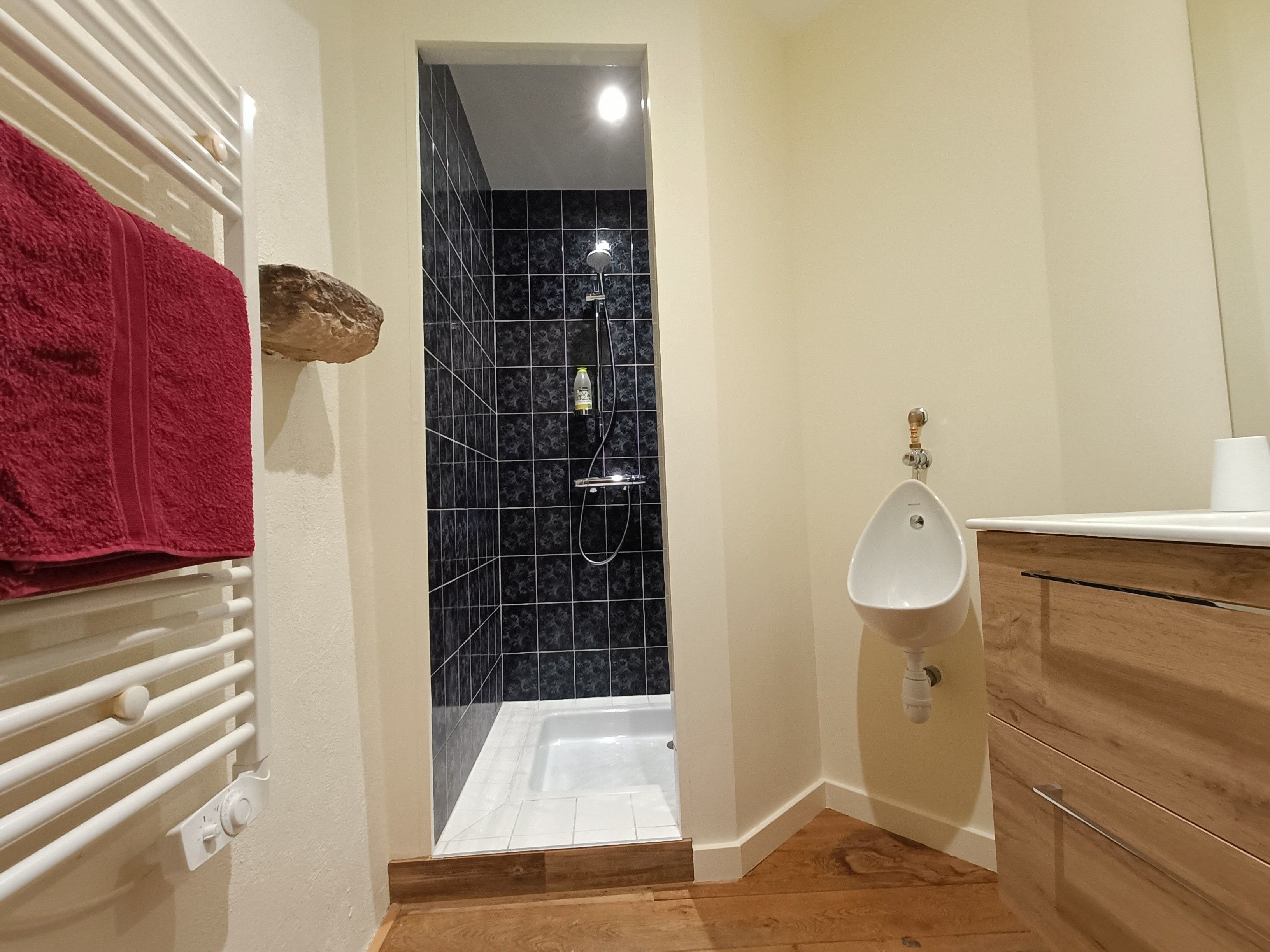 Une salle de bain avec une baignoire blanche, un urinoir blanc, un meuble-lavabo en bois et une serviette rouge posée sur un radiateur.
