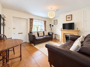 A living room with a fireplace, a television, a sofa, a dining table, and a chandelier. - Pam's Cottage: A Cosy Cotswold Home in Winchcombe (Winchcombe)