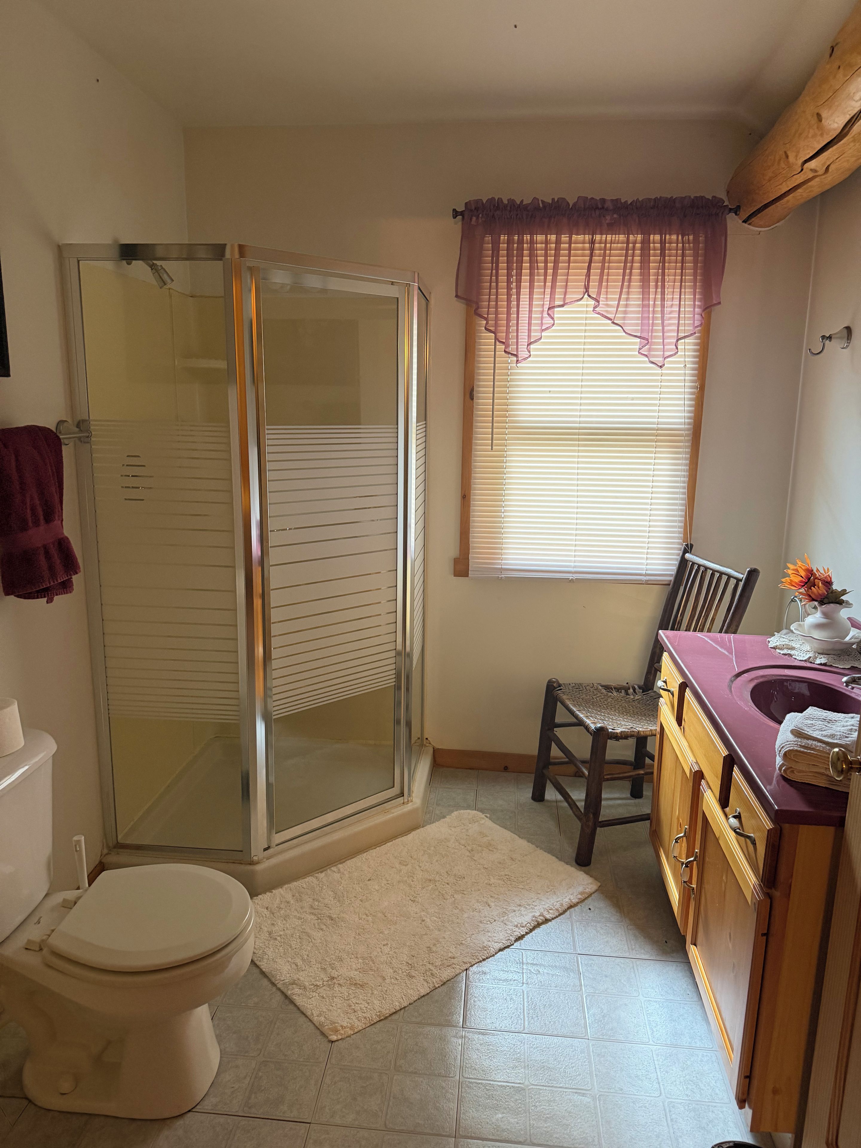A bathroom with a glass shower enclosure, a toilet, a sink with a vanity, and a window with blinds.
