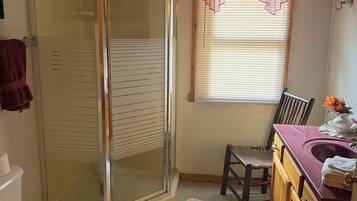 A bathroom with a glass shower enclosure, a toilet, a sink with a vanity, and a window with blinds.