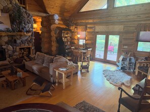 A cozy log cabin interior with a stone fireplace, a wooden dining area, and a large window overlooking a deck.