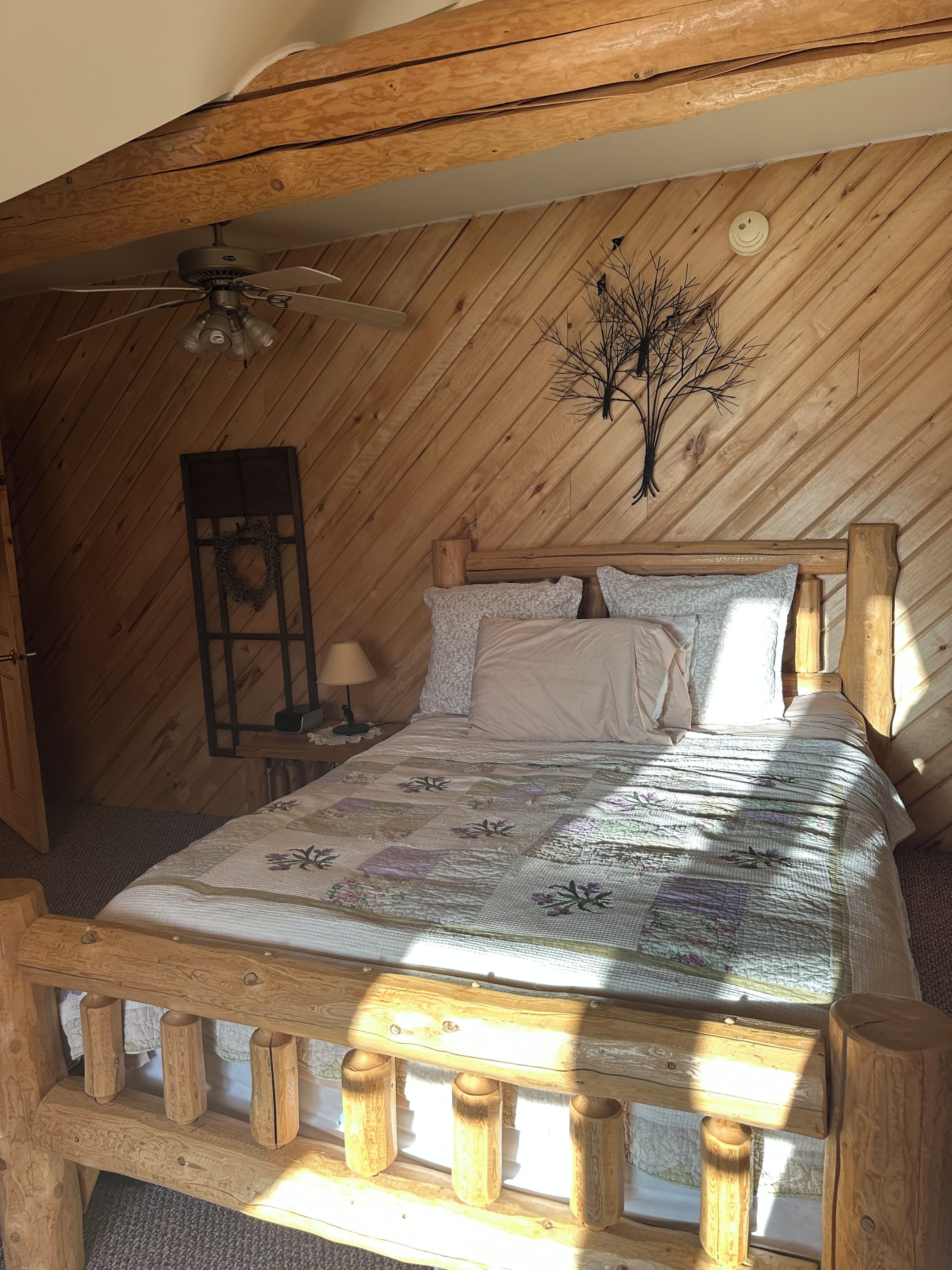 A wooden bed with a quilt in a room with sloped wooden walls and a ceiling fan.