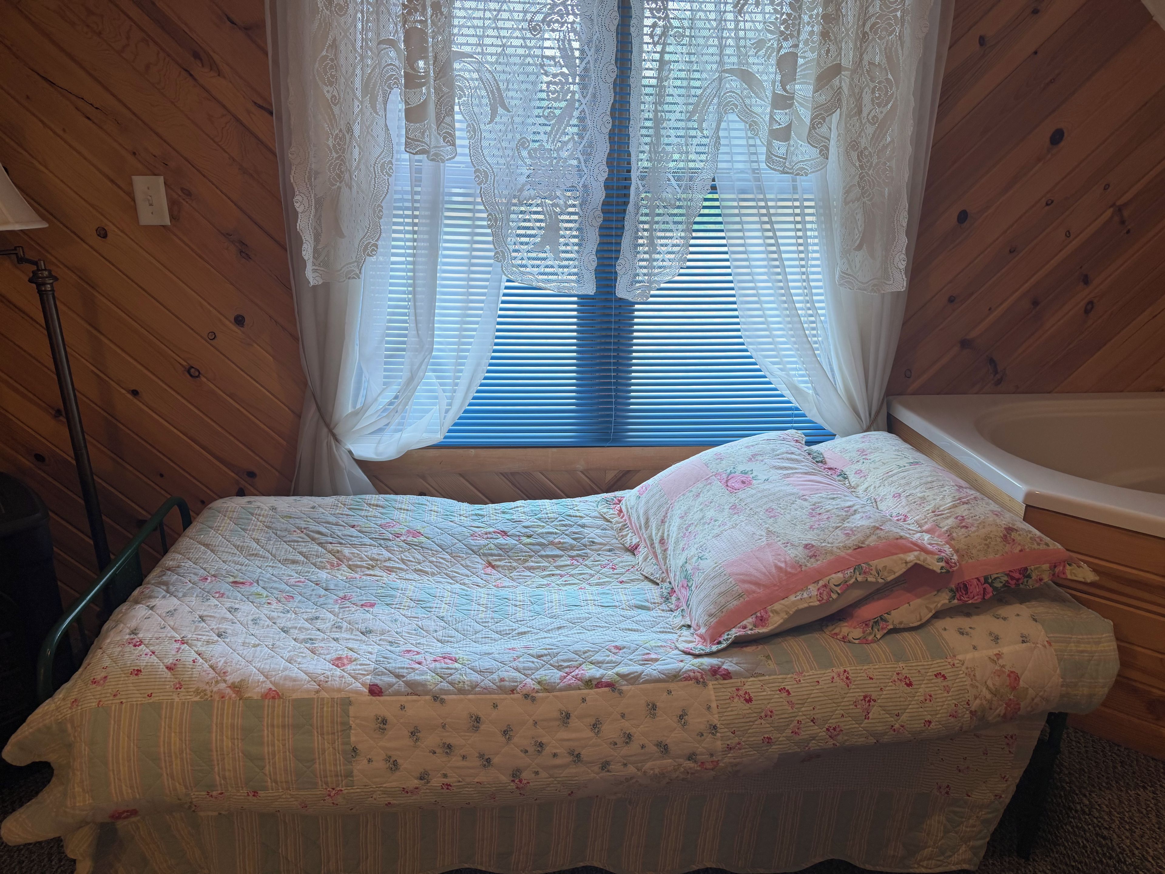 A bedroom with a bed, a window with curtains, a lamp, and a bathtub.