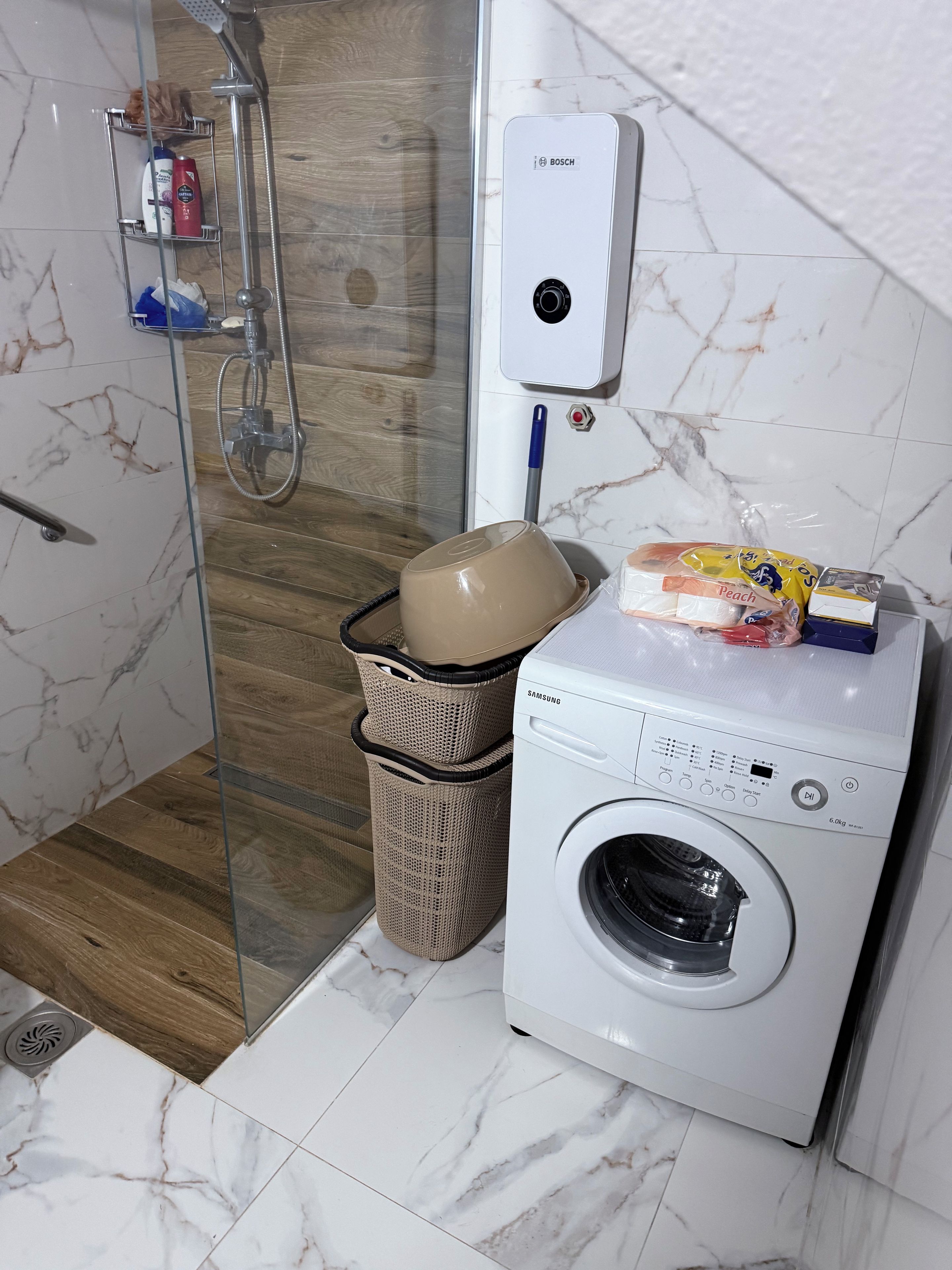 Casa de banho com cabine de duche em vidro, secador de mãos Bosch, máquina de lavar roupa Samsung e cesto de vime.