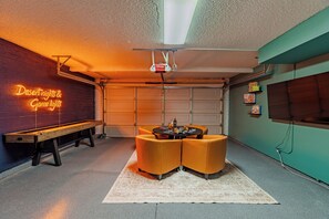 Uma garagem com letreiro de neon, projetor no teto, mesa e cadeiras, e um tapete.