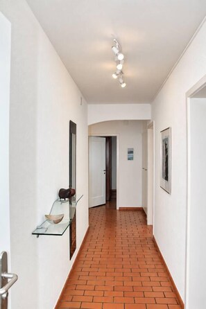 A hallway with a brick floor, white walls, and a glass shelf with a bowl. There is a door at the end of the hallway. - Queen-Size Bed / Private Balcony / Central - Ghost 12 (Zürich)