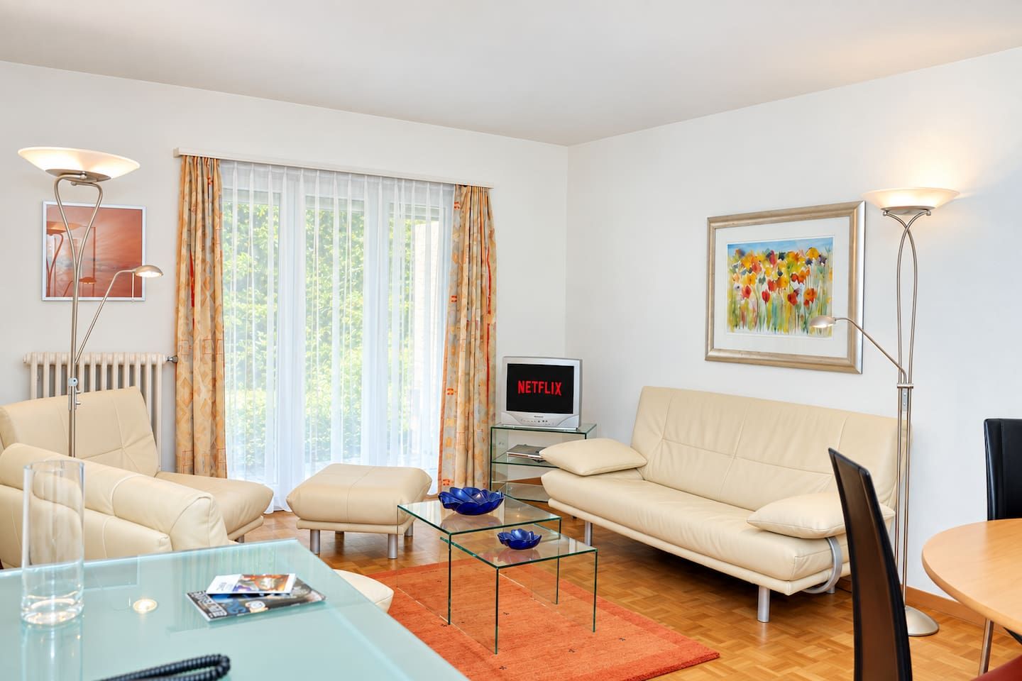 A modern living room with a sofa, armchair, glass coffee table, and a television displaying 'NETFLIX'.