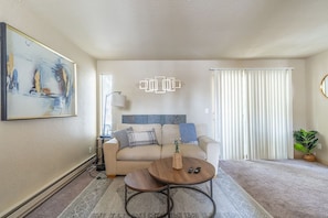 A living room with a beige sofa, a coffee table, a painting on the wall, and a window with blinds. - Centrally Located 1 Bedroom (Seattle)