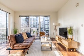 Ein modernes Wohnzimmer mit Ledersofa, Couchtisch, Fernseher auf Holzfuß und Blick auf eine Stadtlandschaft durch große Fenster.