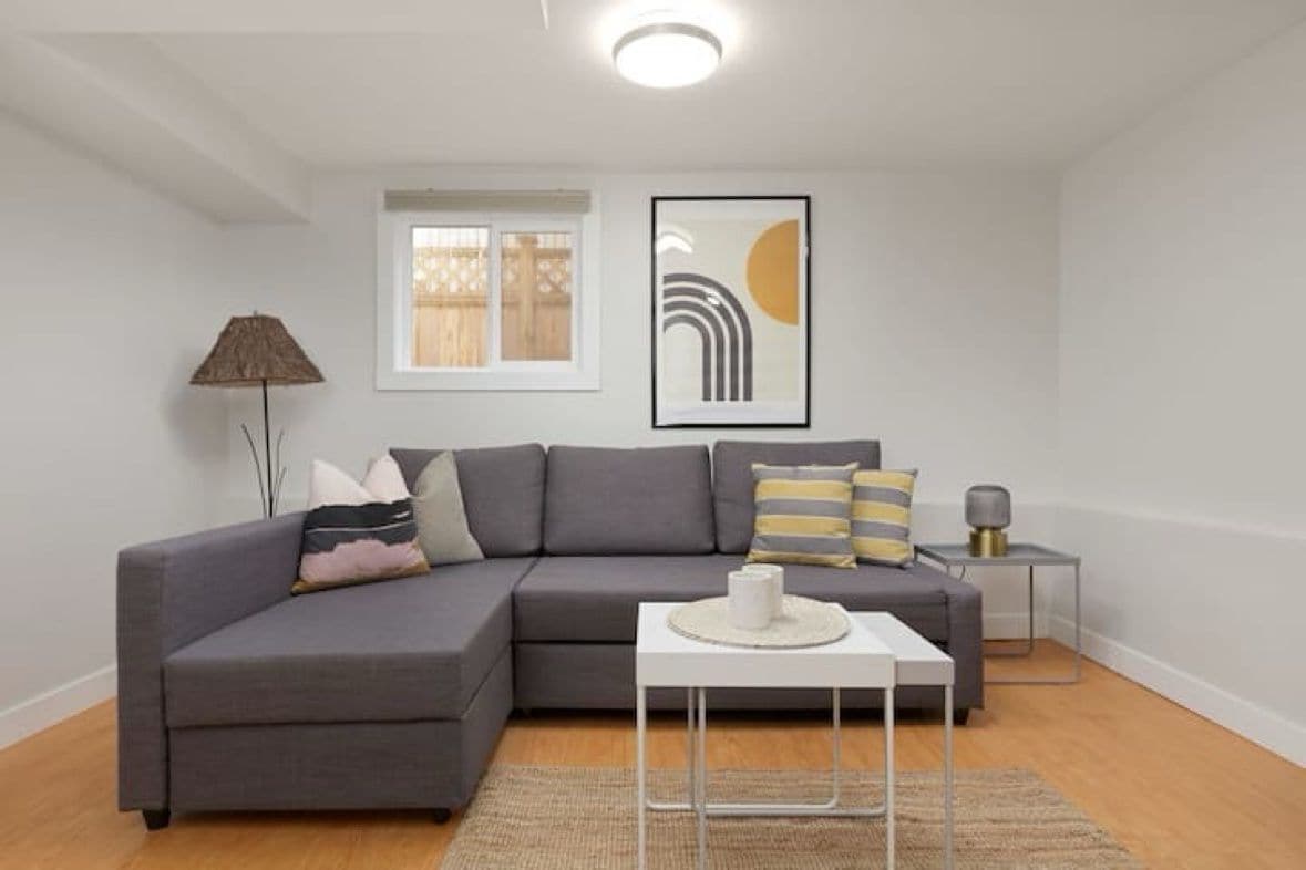 Un salon moderne avec un canapé d’angle gris, une petite table d’appoint blanche, une œuvre d’art abstraite encadrée et une lampe sur pied.