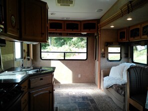 A compact RV interior with a kitchenette, a seating area with a blanket, and a window providing a view of greenery.