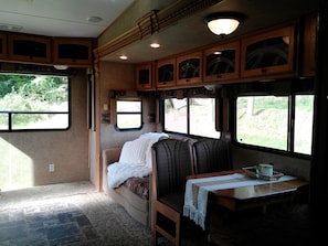 A cozy RV interior with a seating area, a table with a cup, and windows providing views of greenery.