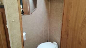 A small, enclosed bathroom with a toilet, a mirror, and a wooden shelf.