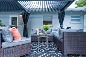 An outdoor seating area with a sofa, a round table, and a decorative wall with shells. - Charlotte Ave Coastal Retreat Private Pool Fire Pit Backyard Lounge (Sarasota)