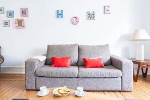 Ein graues Sofa mit roten Kissen, ein Couchtisch aus Holz mit einer Teller Kekse und einer Tasse, und eine Wand mit Buchstaben, die das Wort 'HOME' bilden.