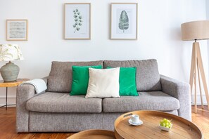 Ein Wohnzimmer mit grauem Sofa, grünen und weißen Kissen, einem Holztisch mit einer Tasse und Trauben sowie gerahmten Pflanzenprints an der Wand.