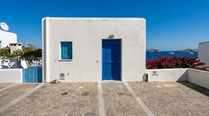 Ein weißes Gebäude mit einer blauen Tür und einer Satellitenschüssel, eine gepflasterte Fläche und ein freier Blick auf das Meer und Boote im Hintergrund.