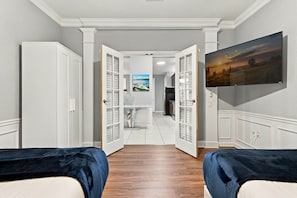 A hotel room with a wooden floor, a bed with a blue cover, a TV mounted on the wall, and a painting of a landscape. - 3BR close to Convention Center and Theme Parks (Orlando)