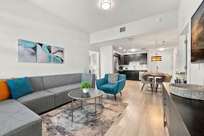 A modern living room with a grey sofa, a blue armchair, and a coffee table. A kitchen is visible in the background with dark cabinets and stainless steel appliances. - Spacious 3BR condo, Perfect Orlando Home Base (Orlando)