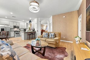 Ein modernes Wohnzimmer mit einem beigefarbenen Sofa, einem Glastisch und einer Küche im Hintergrund.