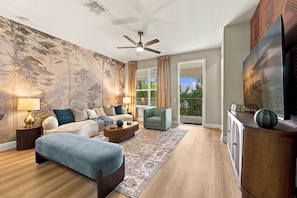 A modern living room with a sofa set, a coffee table, a floor lamp, a ceiling fan, a large window with curtains, and a television on a cabinet. - Luxury 3BR, Modern Design, Lake Views, Vista Cay (Orlando)