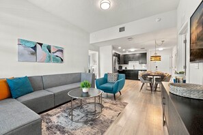 Ein modernes Wohnzimmer mit einem grauen Sofa, einem blauen Sessel und einem Couchtisch. Im Hintergrund ist eine Küche mit dunklen Schränken und Edelstahlgeräten zu sehen.