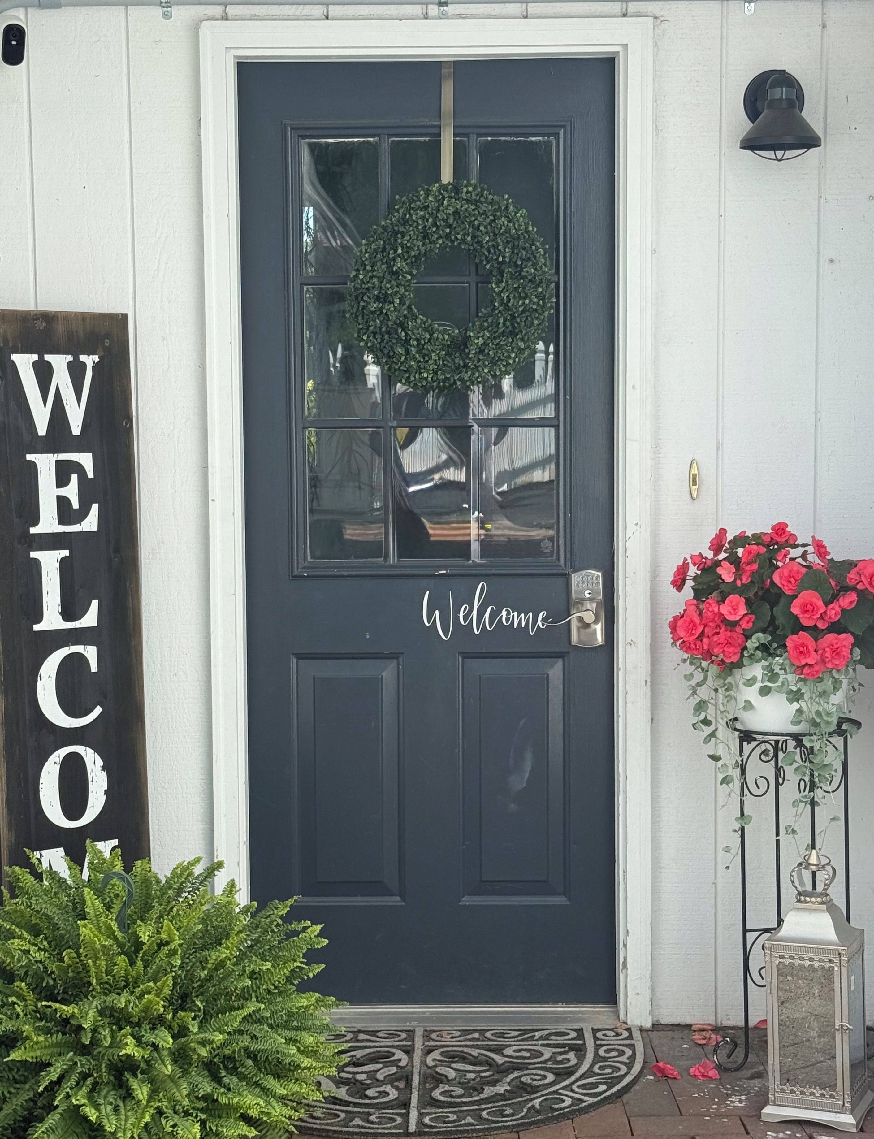 Eine schwarze Tür mit einem Kranz und einem 'WELCOME'-Schild, ein Topf mit roten Blüten und ein Willkommen-Schild an der Wand.