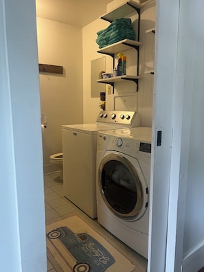 A laundry room with a front-loading washing machine, a dryer, and shelves above holding folded clothes and laundry supplies. - Charming 4-bedroom house with POOL in peaceful New Columbia (New Columbia)