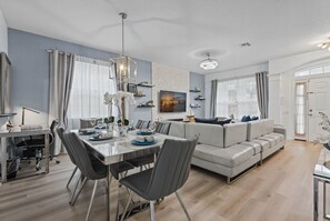 A modern living room with a dining area, grey sofas, and a flat-screen TV mounted on the wall. - Chic 6BR, Pool, Themed Rooms, Near Disney (Four Corners)
