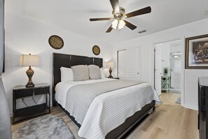 A bedroom with a bed, two bedside tables, a ceiling fan, and a door leading to a bathroom. - Chic 6BR, Pool, Themed Rooms, Near Disney (Four Corners)