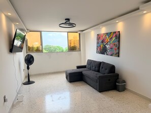 A modern living room with a sectional sofa, a flat-screen TV mounted on the wall, a ceiling fan, and a large window with a city view. - Cozy 3-bedroom en La Zona Rosa de Barranquilla (Barranquilla)