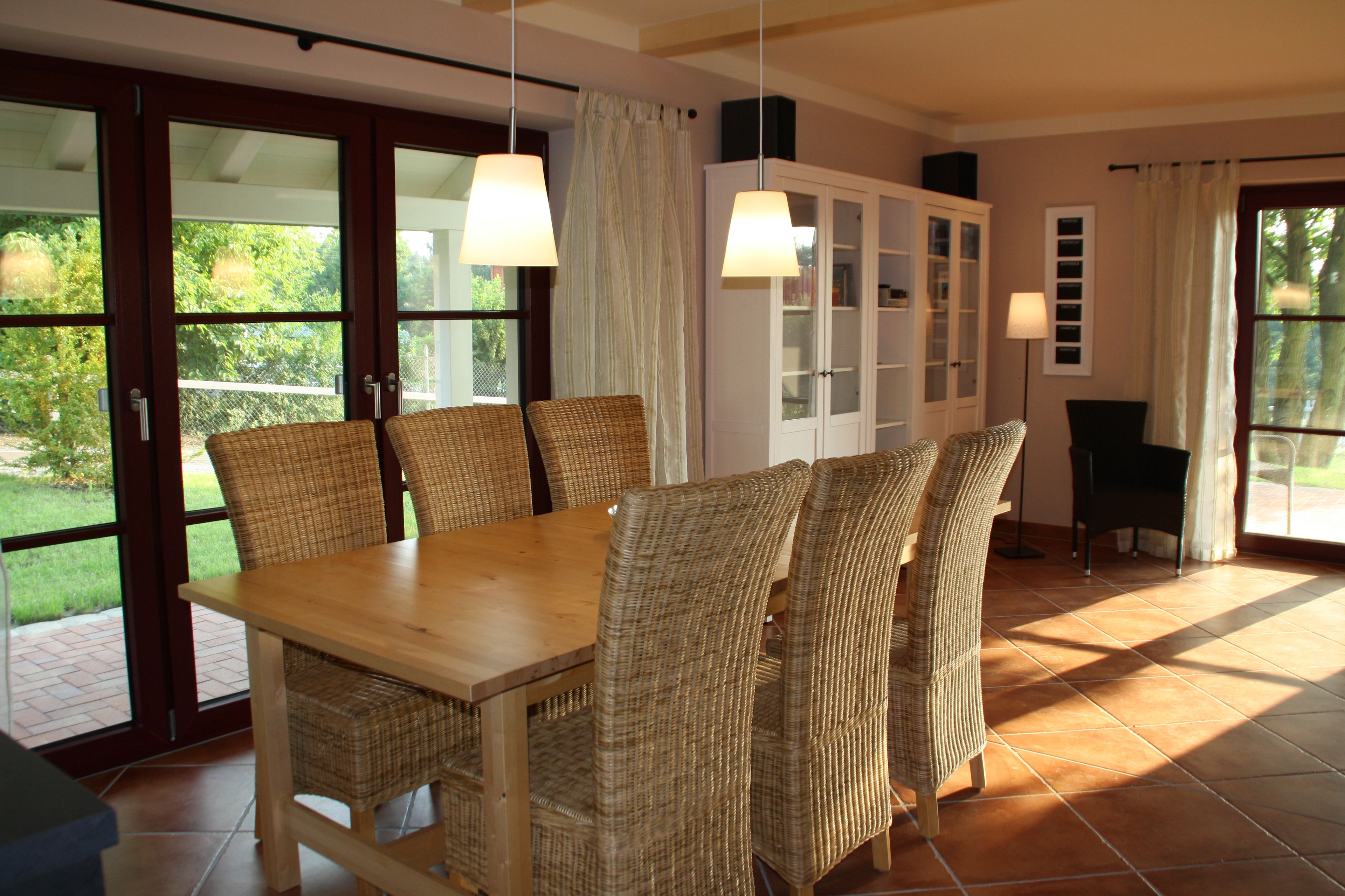 A dining area with a wooden table and wicker chairs, a cabinet with glass doors, and a view of greenery through large sliding doors.