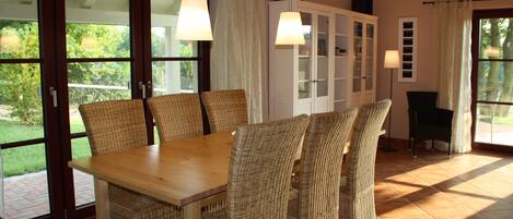 A dining area with a wooden table and wicker chairs, a cabinet with glass doors, and a view of greenery through large sliding doors.