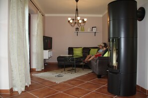 A living room with a sofa, a television, a fireplace, and a chandelier. - Cozy vacation home with a fireplace on a large garden lot, with a view of Lilienstein (Rathmannsdorf)