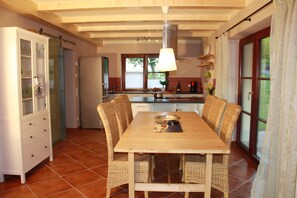 A dining area with a wooden table and wicker chairs, a kitchen in the background, and a view of the outdoors through a window. - Cozy vacation home with a fireplace in a large garden, with a view of Lilienstein   (Rathmannsdorf)