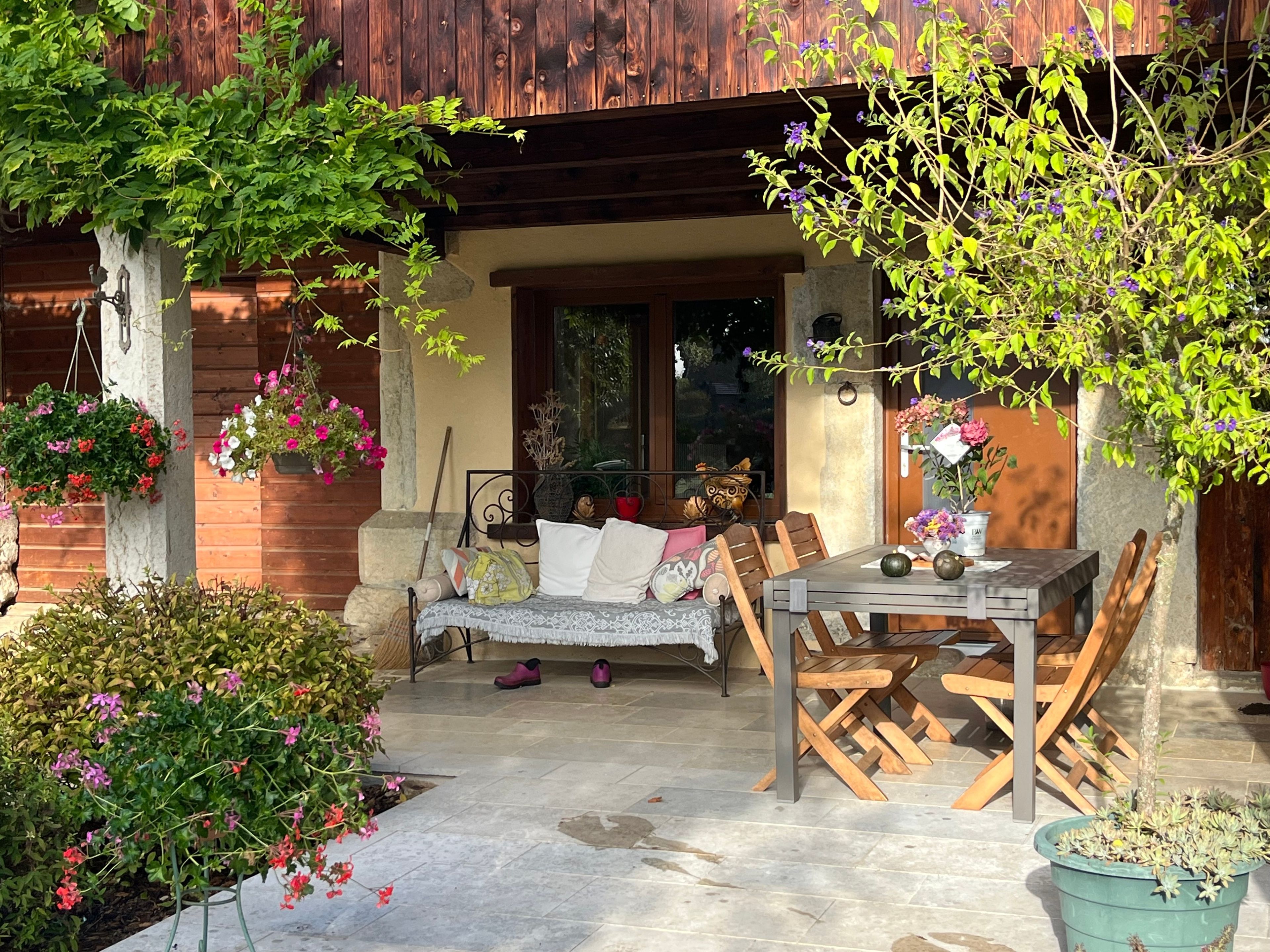 A patio with a wooden table and chairs, a bench with cushions, and hanging flower baskets.