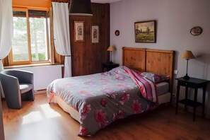 Ein Schlafzimmer mit Bett, Sessel, Nachttisch, Lampe und einem Fenster mit Vorhängen.